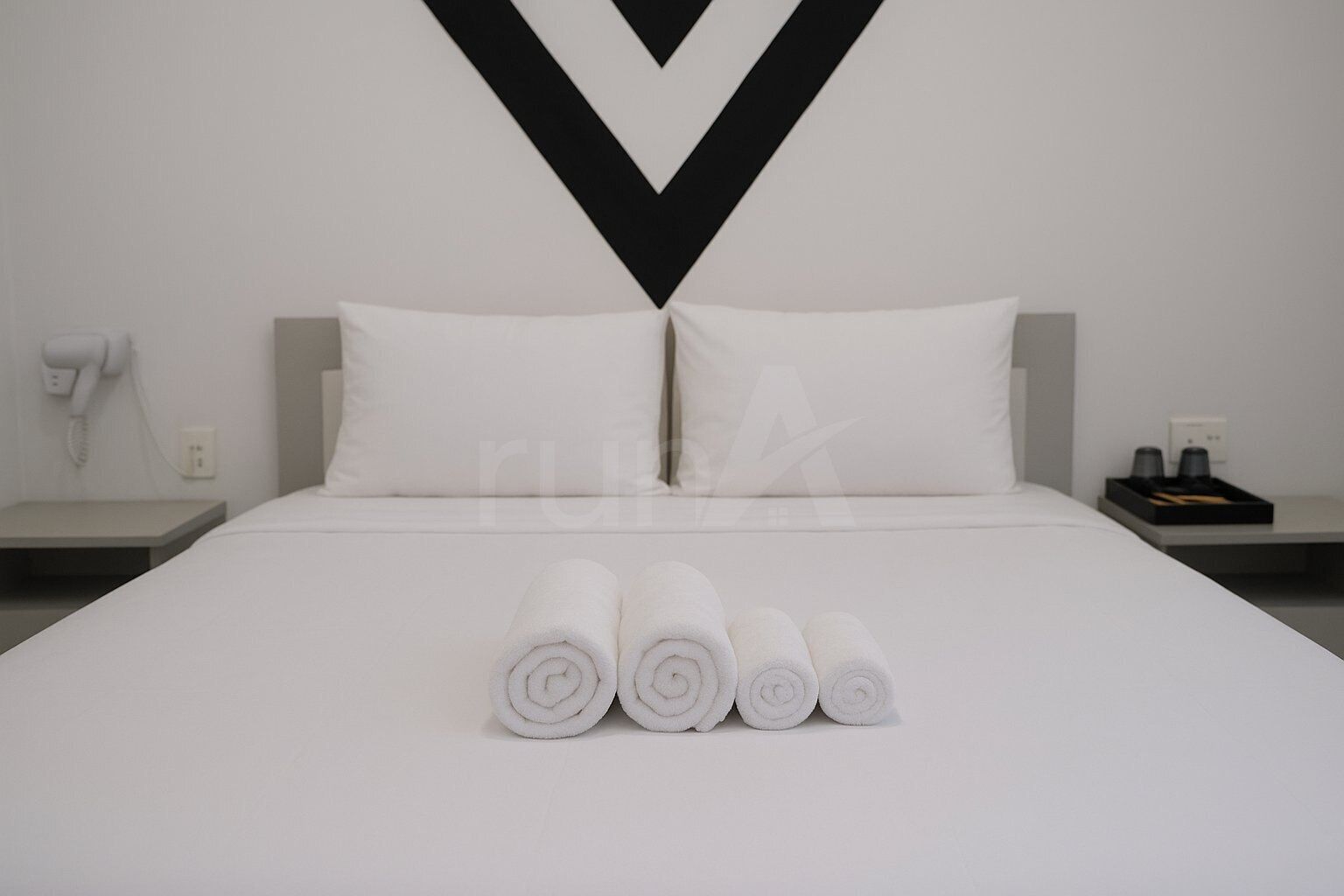 A neatly made bed with rolled towels in front, a modern wall design, and a small bedside table with a phone and a coffee maker.