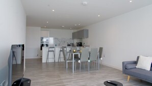 A modern living room with a grey sofa, a small black table, and a TV. A kitchen with a dining area is visible in the background. - Stylish 3BR | 3 Min to Southfields Station (Greater London)