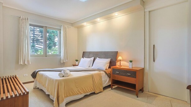 A bedroom with a bed, bedside table, lamp, and a view of trees through the window. - Apart w Beautiful View in Campos do Jordao Lvr0003 (Campos do Jordão)