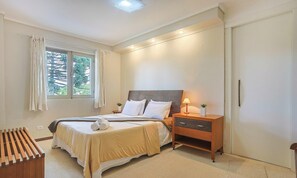 Un dormitorio con cama, mesita de noche, lámpara y vistas a árboles por la ventana.
