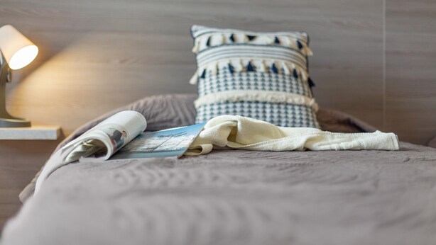 Un lit bien fait, avec un oreiller à motifs, un magazine enroulé et une lampe sur une table de chevet.