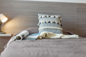 Una cama bien hecha con un almohadón estampado, una revista enrollada y una lámpara sobre una mesita de noche.