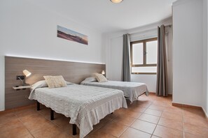 A room with two beds, a nightstand with a lamp, a window with curtains, and a wall-mounted picture.