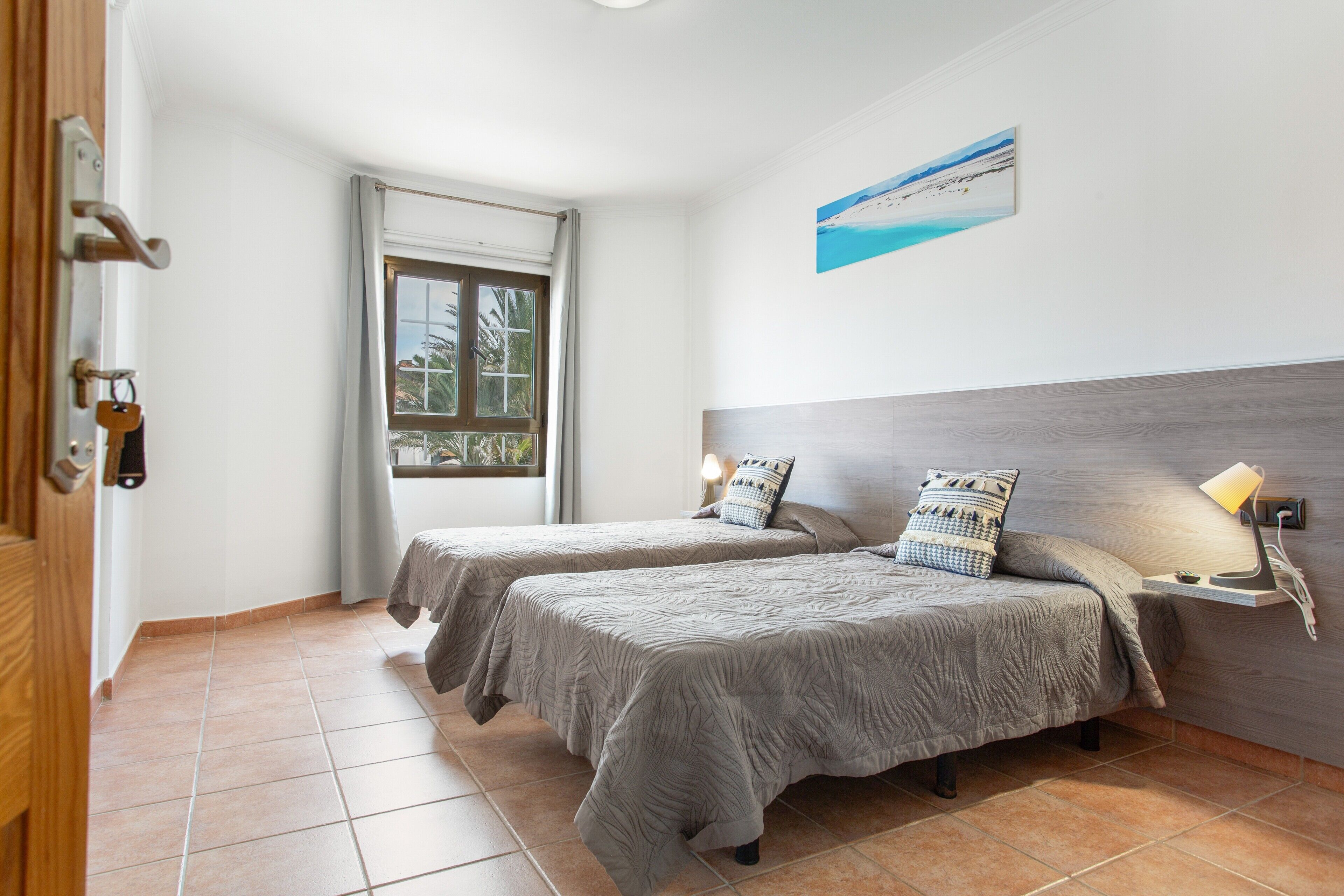 A hotel room with two beds, a window with curtains, a wall-mounted painting, and a bedside table with a lamp.