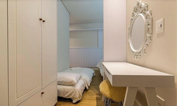 Un dormitorio con un armario blanco, una cama con ropa de cama blanca, una tocador con espejo y un taburete pequeño.