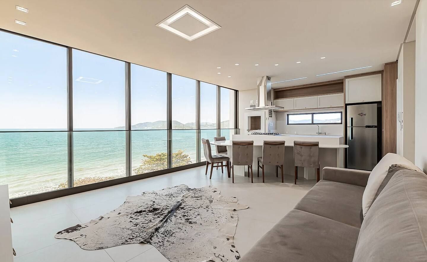 A modern kitchen with large windows overlooking a body of water, a dining area with chairs, and a large rug on the floor.