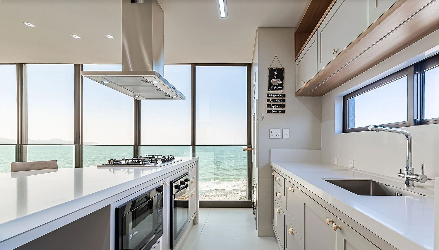 A modern kitchen with a large island, built-in oven, and a view of the sea.