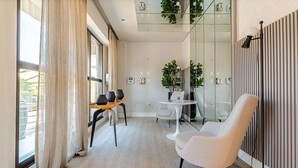 Una habitación de hotel moderna con un ventanal grande, una zona de estar con una mesa y sillas, un sillón blanco y una planta en maceta.