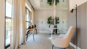 Quarto de hotel moderno com uma janela grande, área de estar com mesa e cadeiras, uma poltrona branca e um vaso de planta.