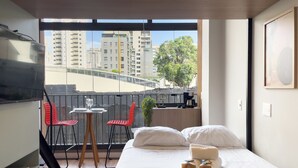 Hotelkamer met een bed, een kleintje tafeltje met stoelen, een koffieapparaat en uitzicht op de stad door grote ramen.