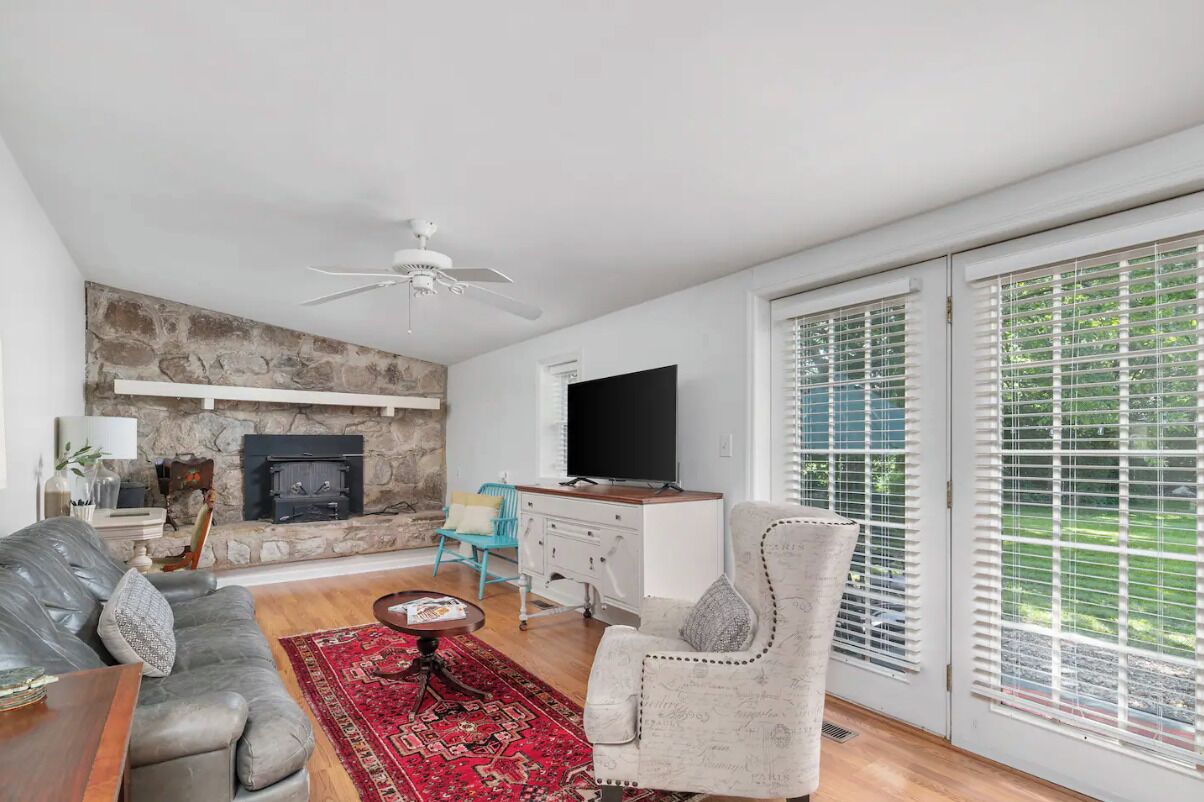 A living room with a stone fireplace, a flat-screen TV, a sofa, a chair, and a large window with blinds.