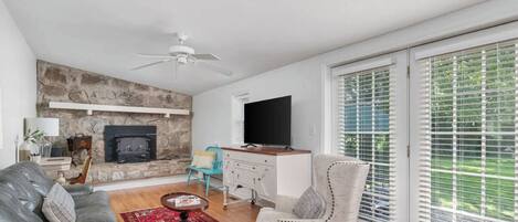 A living room with a stone fireplace, a flat-screen TV, a sofa, a chair, and a large window with blinds.