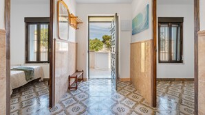 A hallway with patterned tiles, a bed with a green blanket, a wooden bench, and a view through an open door to a balcony.