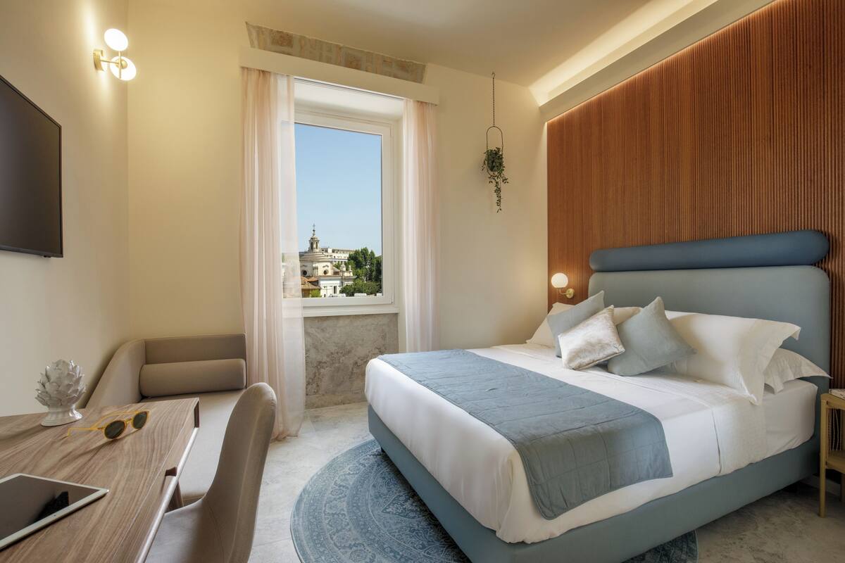 Ein modernes Hotelzimmer mit einem großen Bett, einem Schreibtisch mit einem Tablet, einem Sessel, einem Fernseher und Blick auf eine Stadtlandschaft durch ein Fenster.