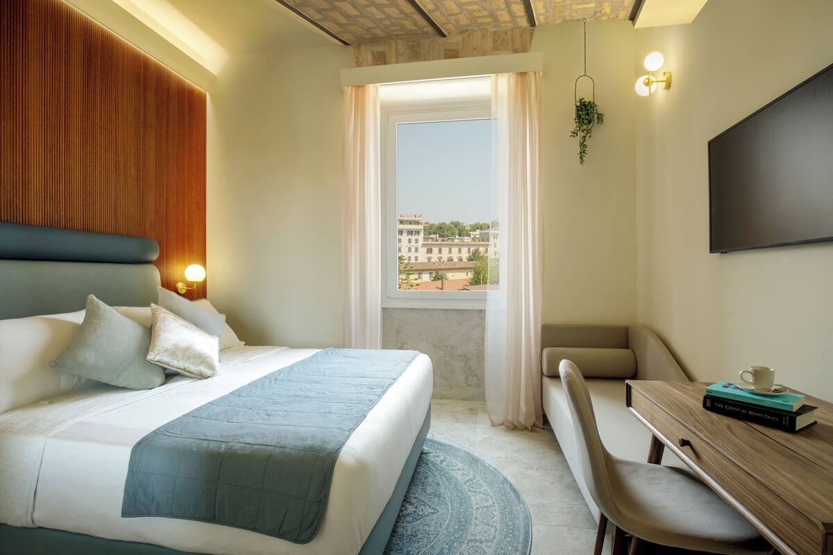 Ein Hotelzimmer mit Bett, Schreibtisch, Stuhl, Fernseher und Fenster mit Blick auf Gebäude.