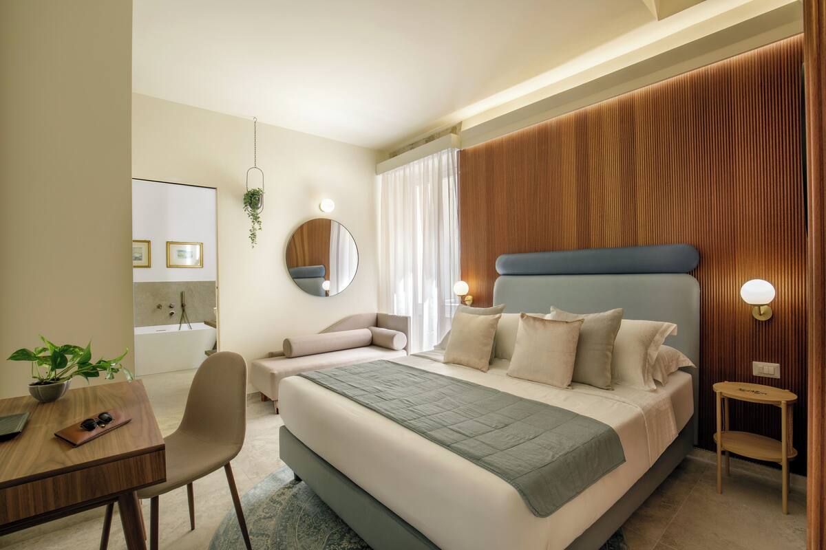Ein modernes Hotelzimmer mit einem großen Bett, einem Schreibtisch und einem Sessel. Es gibt einen Spiegel, eine Pflanze und einen kleinen Tisch mit einer Lampe.