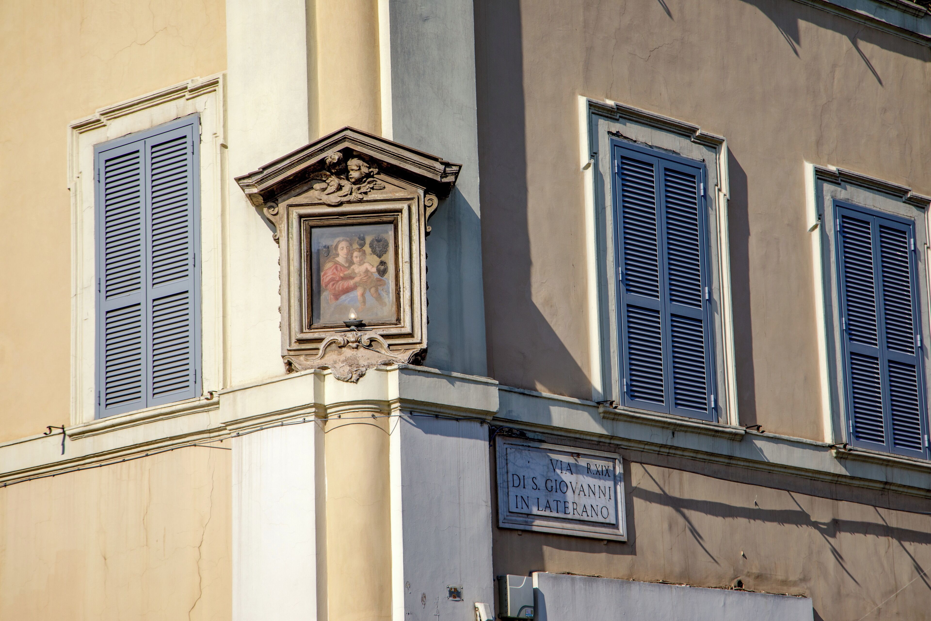 A building corner with three windows, each with blue shutters. A decorative frame with a painting is above the central window. A sign reads "Via RMX D.S. Giovanni in Laterano."