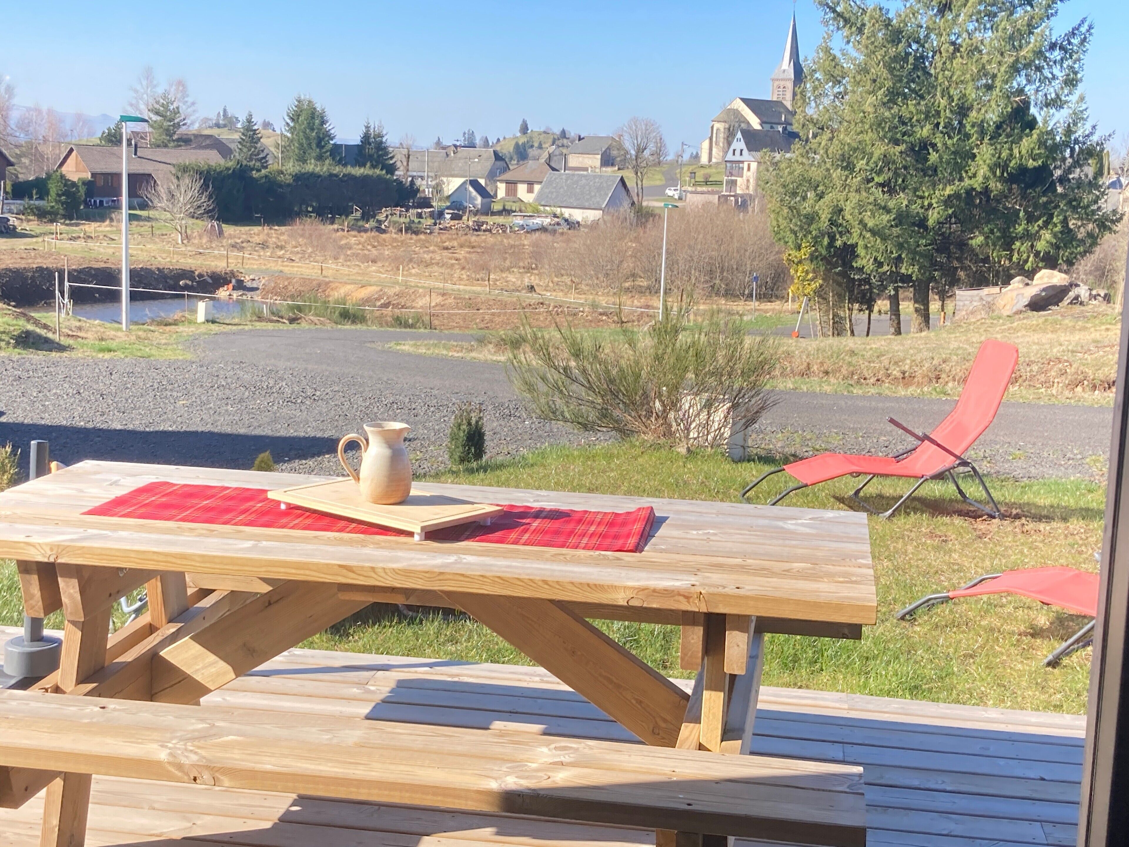 Een houten picknicktafel met een waterkan en een rode placemat, een rode loungestoel en een uitzicht op een dorp met huizen en een kerk op de achtergrond.