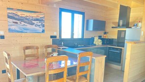 A wooden interior with a dining table, chairs, a kitchen area, and a painting on the wall.