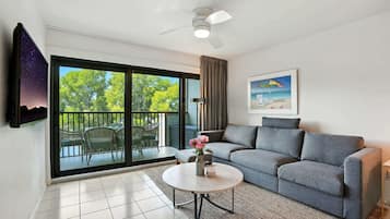A living room with a grey sofa, a round coffee table with a vase and candle, a ceiling fan, and a large sliding glass door leading to an outdoor patio with chairs and a table.