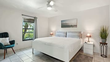 A bedroom with a bed, a chair, a nightstand with a lamp, a ceiling fan, a window with a view of greenery, and a painting on the wall.