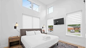A modern bedroom with a large bed, a nightstand, a TV mounted on the wall, and large windows with blinds.