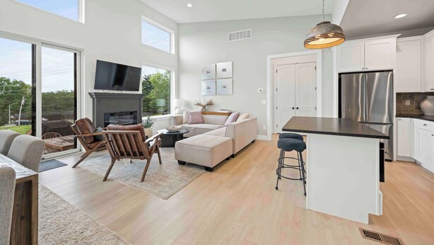 A spacious living area with a large window, a fireplace, a sofa, and a kitchen with white cabinets and a stainless steel refrigerator.