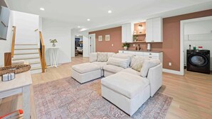 Ein modernes Wohnzimmer mit einem Ecksofa, einem Essbereich mit Tisch und Stühlen, einer Küche mit weißen Schränken sowie einer Waschmaschine.
