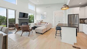 A spacious living area with a large window, a fireplace, a sofa, and a kitchen with white cabinets and a stainless steel refrigerator.