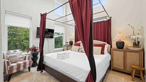 A four-poster canopy bed with red curtains, a television on the wall, a side table with a lamp, and a view of trees through the window.
