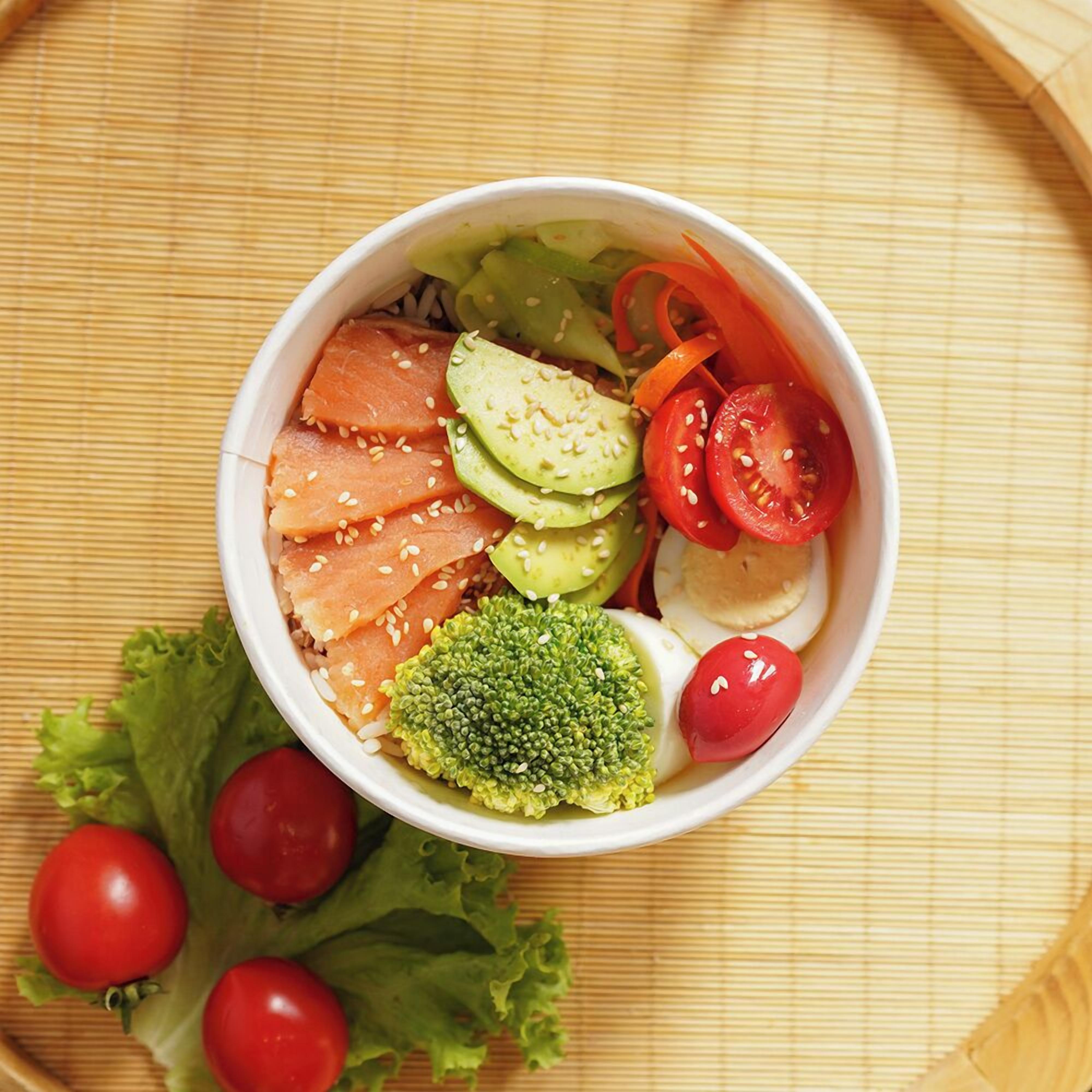 A bowl of sashimi with salmon, avocado, cucumber, and cherry tomatoes on a bamboo mat.