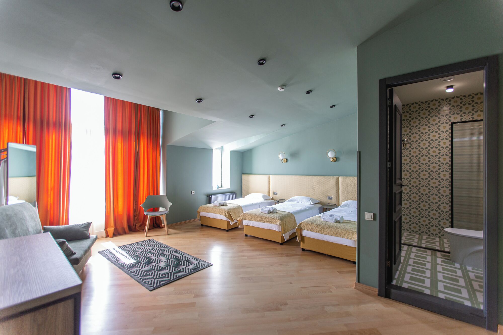 A modern hotel room with two single beds, a sofa, a dining table, and a patterned rug.