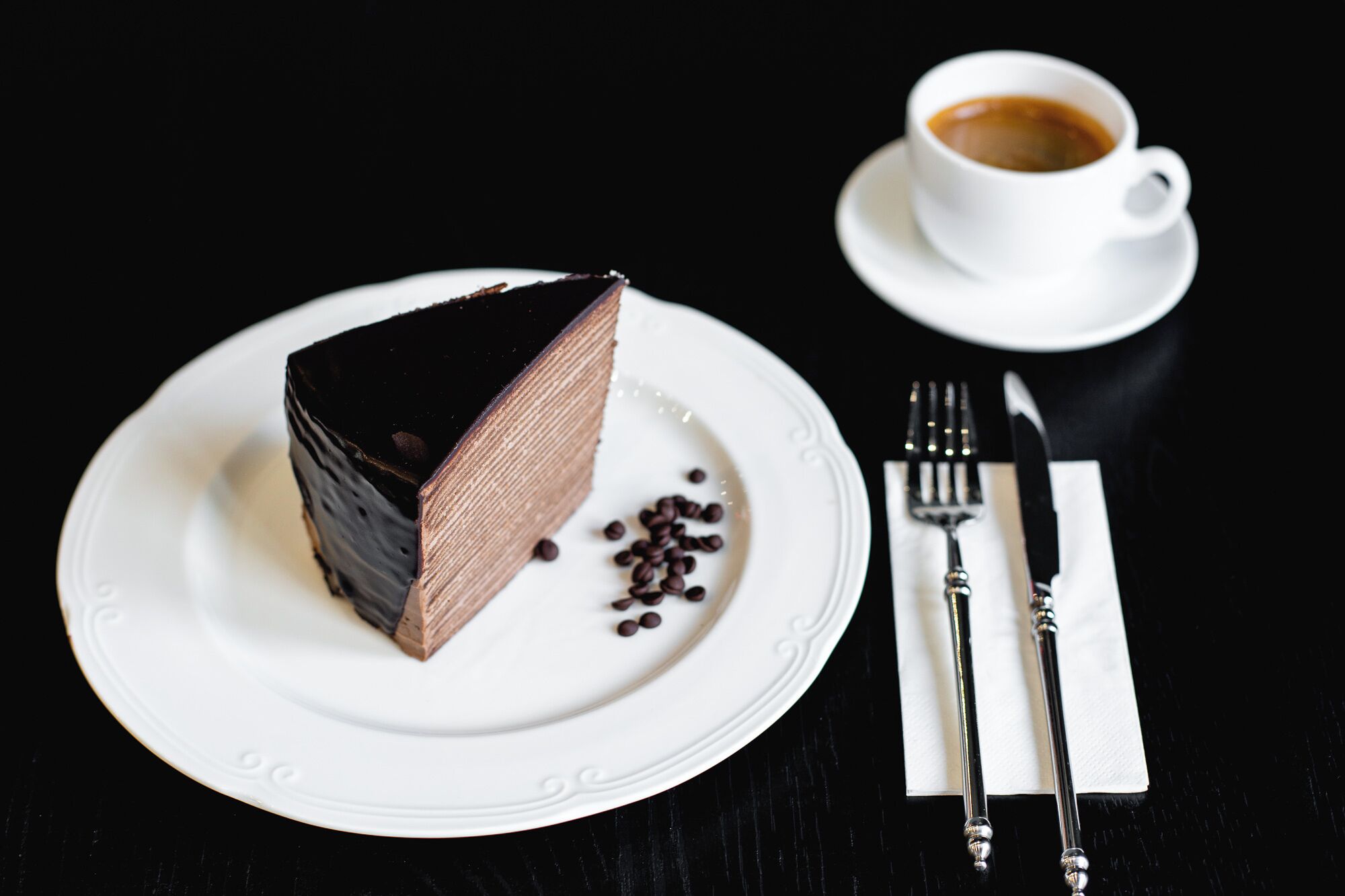 Une part de gâteau au chocolat sur une assiette blanche, accompagnée d’une tasse de café également blanche, ainsi qu’une fourchette et un couteau posés sur une serviette.