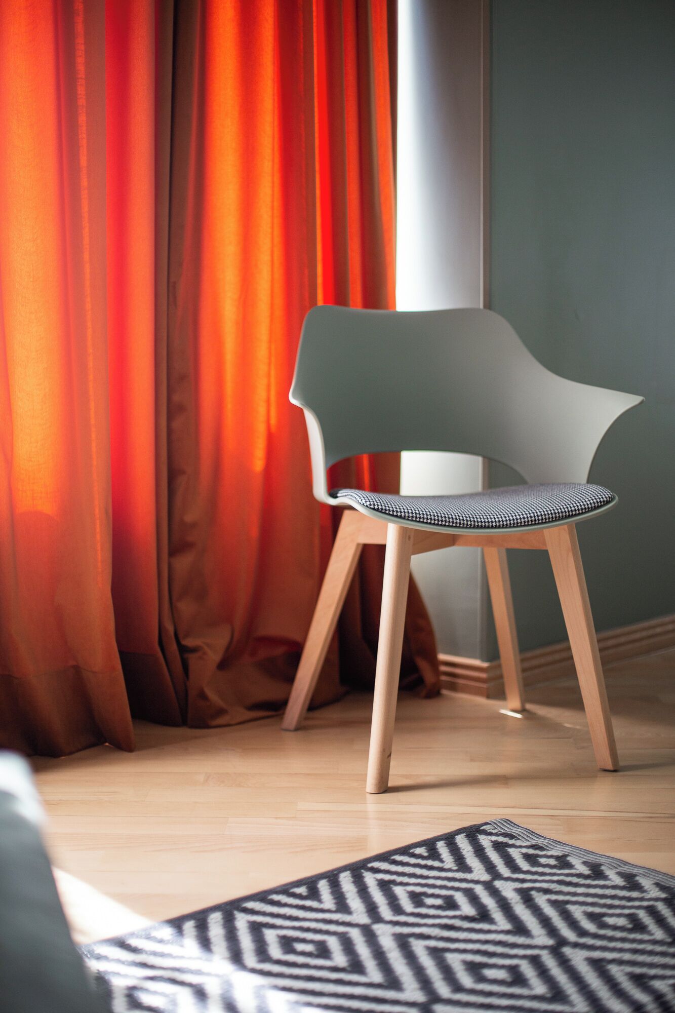 Una habitación moderna con piso de madera, un tapete estampado, una silla verde con asiento tejido y cortinas naranjas.