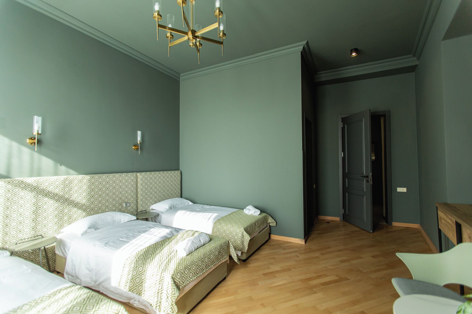 A room with two beds, a wooden floor, a chandelier, and a door leading to another room.