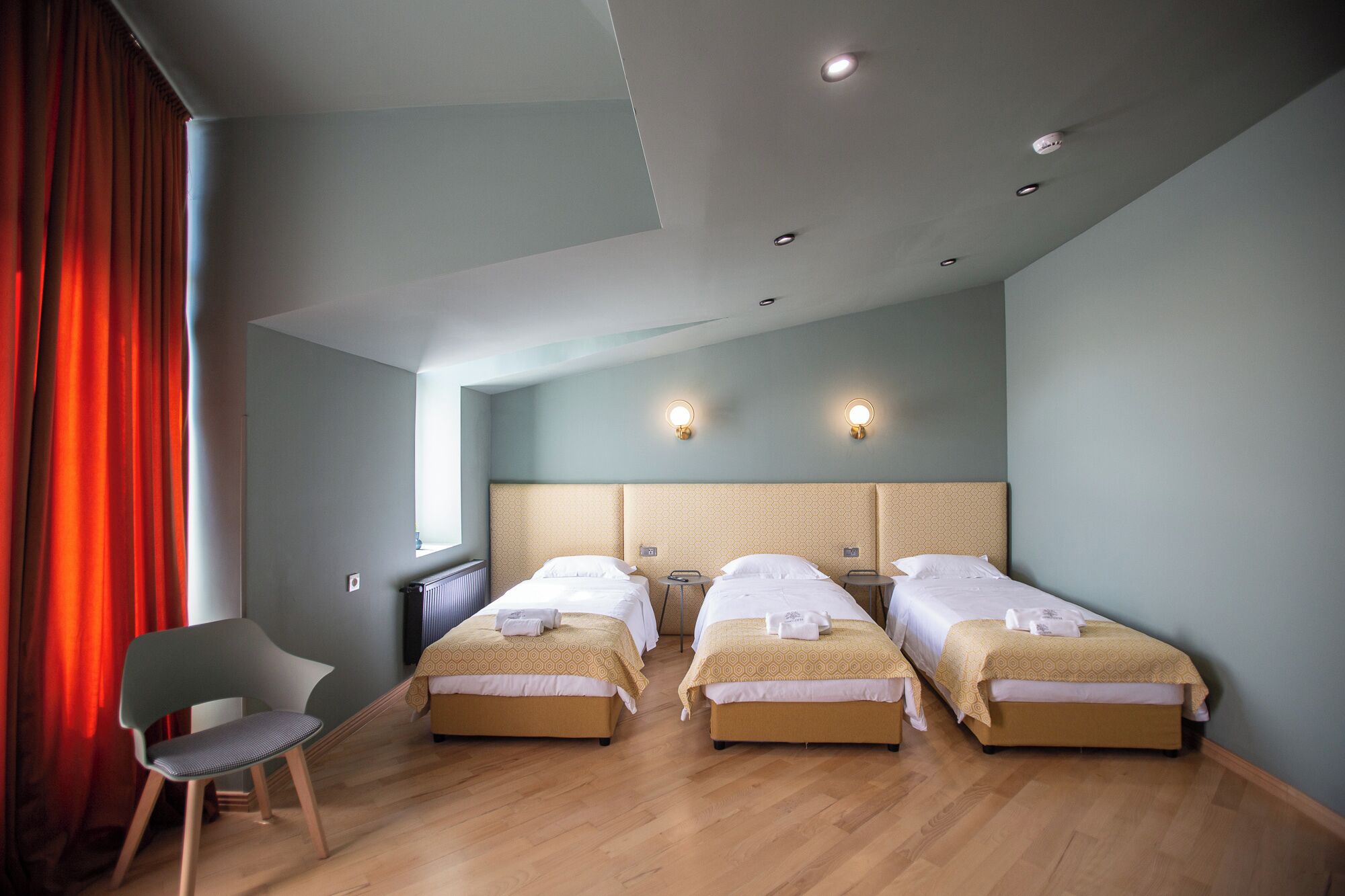 A hotel room with three single beds, a chair, and a window with red curtains.