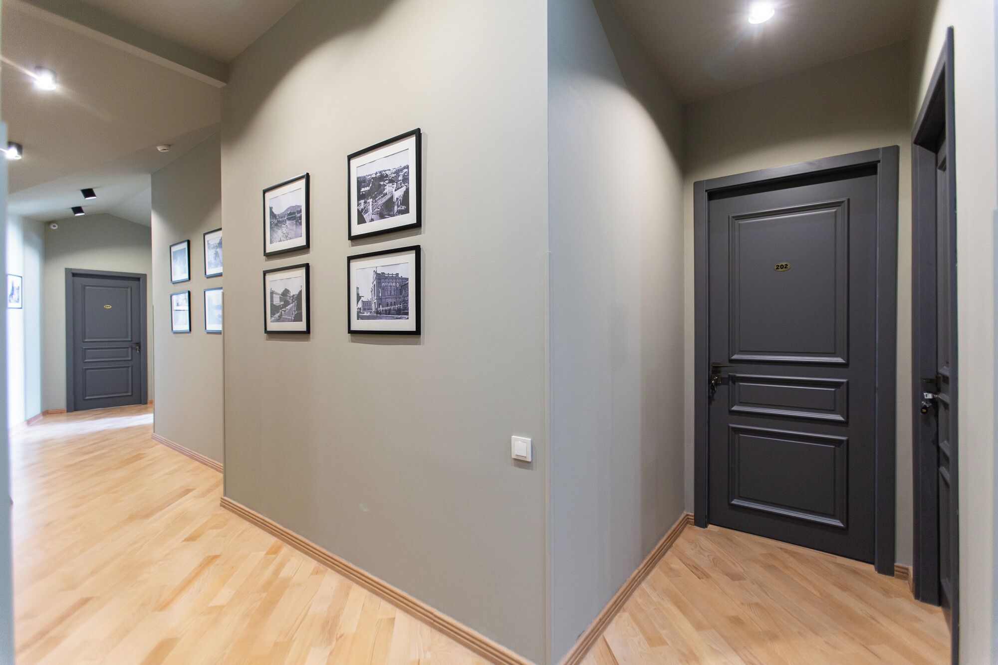 A hallway with a wooden floor, a grey wall with framed pictures, and a dark door with a number plate.