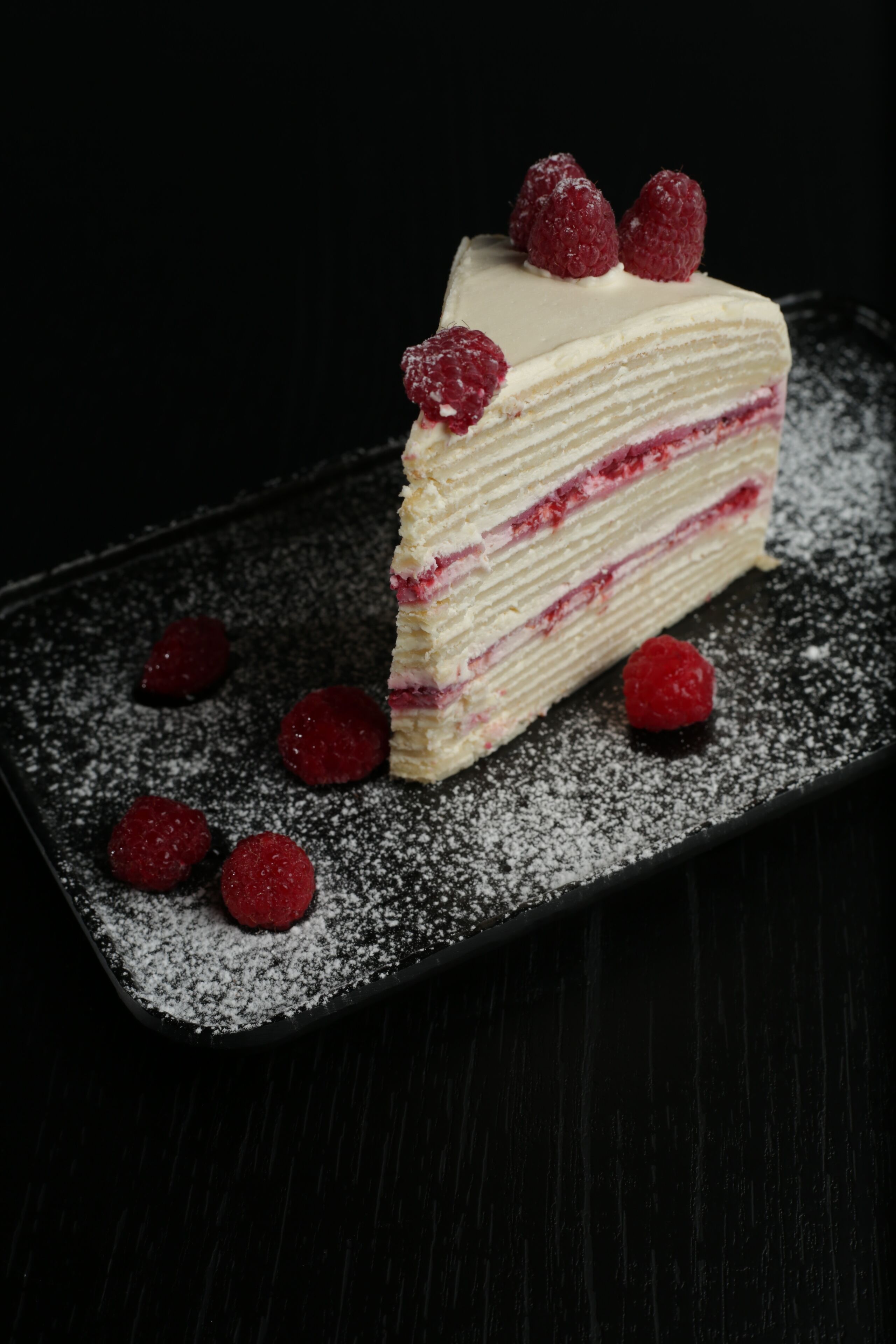 A slice of layered cake with raspberries on a black plate.