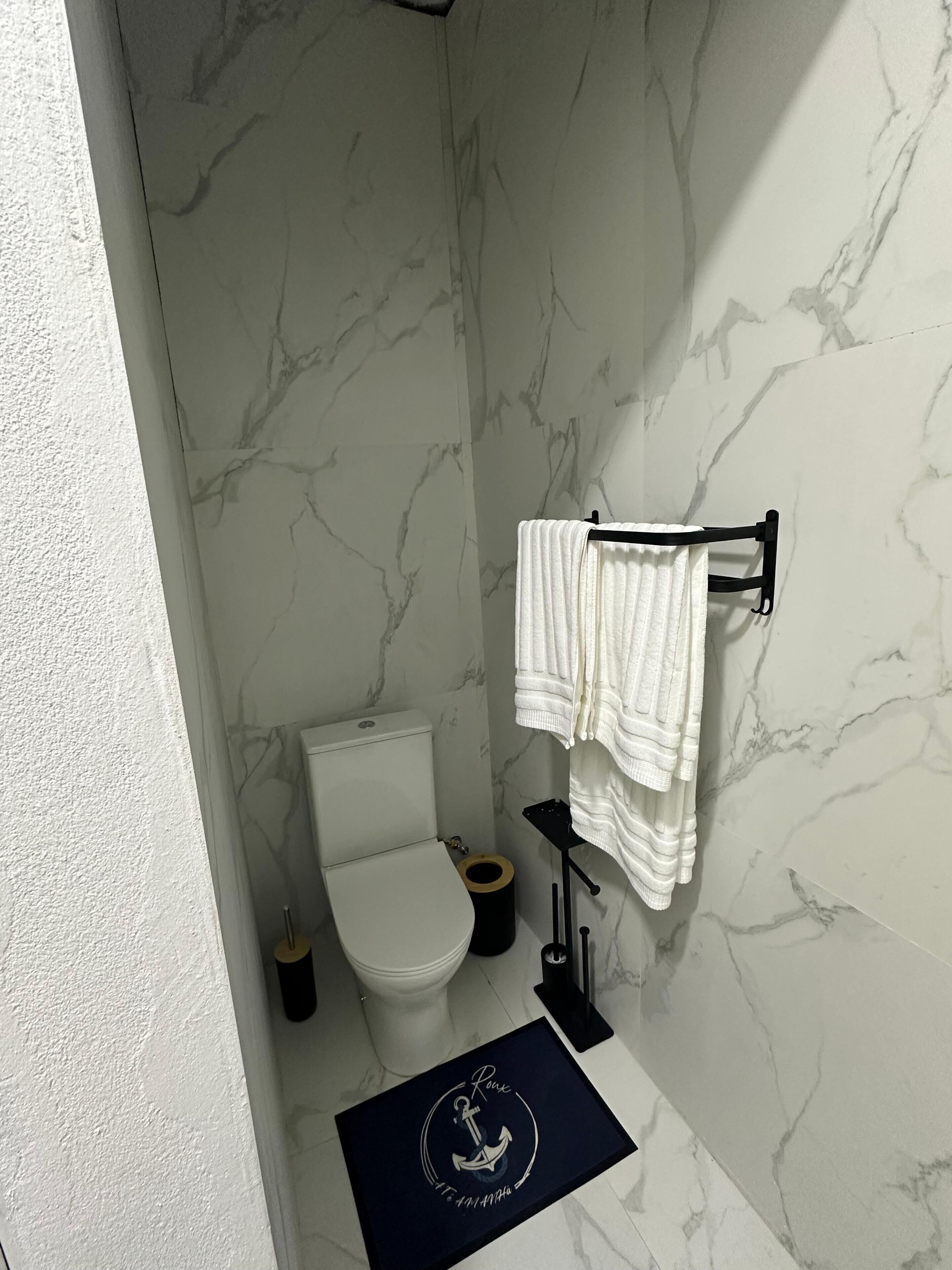 Une salle de bain comprenant des toilettes, un porte-serviettes avec une serviette, un tapis noir orné d’un motif d’ancre et des murs à motifs de marbre.