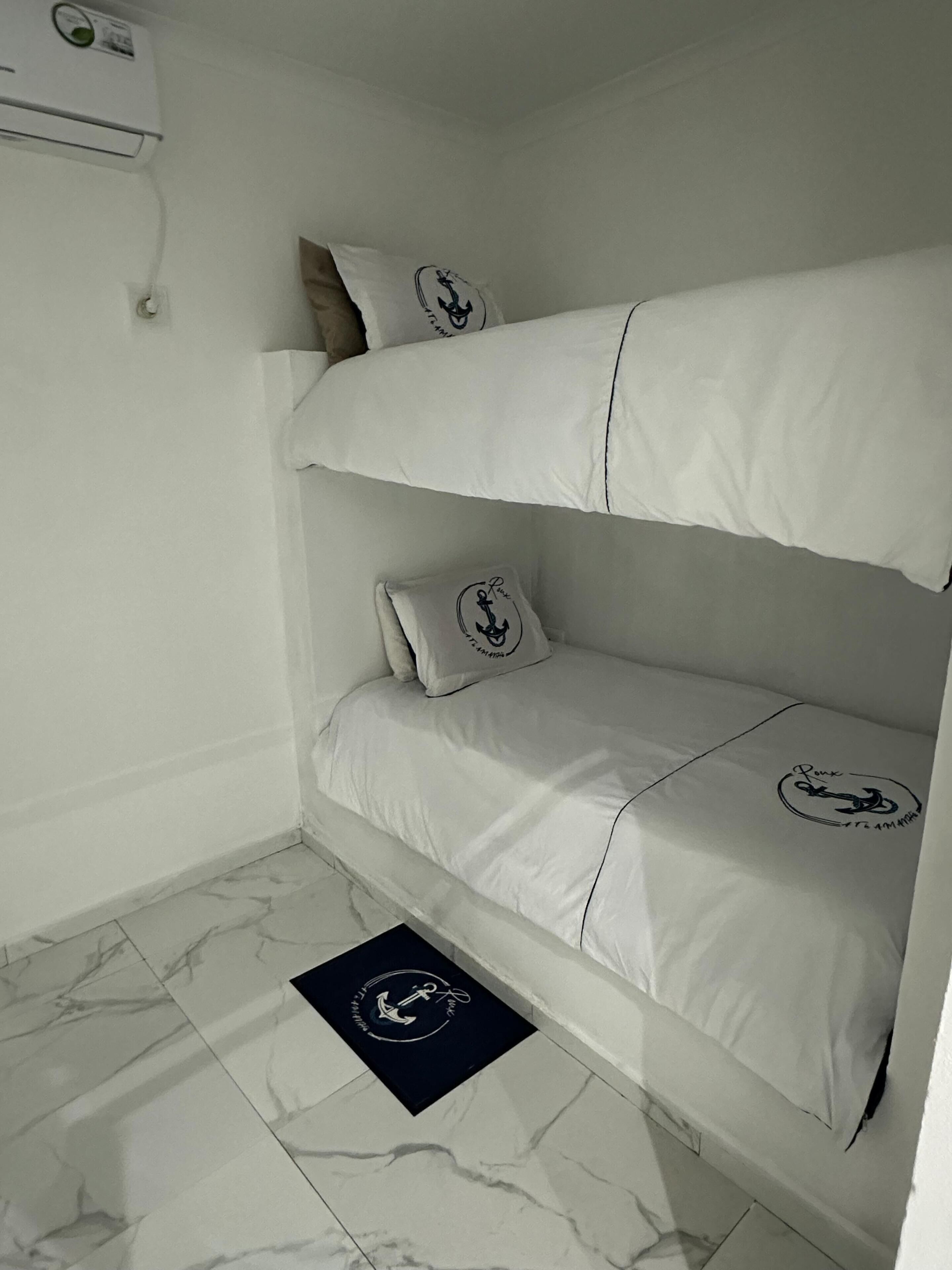 A bunk bed with two pillows, each featuring an anchor logo, in a room with white walls and a marble floor.