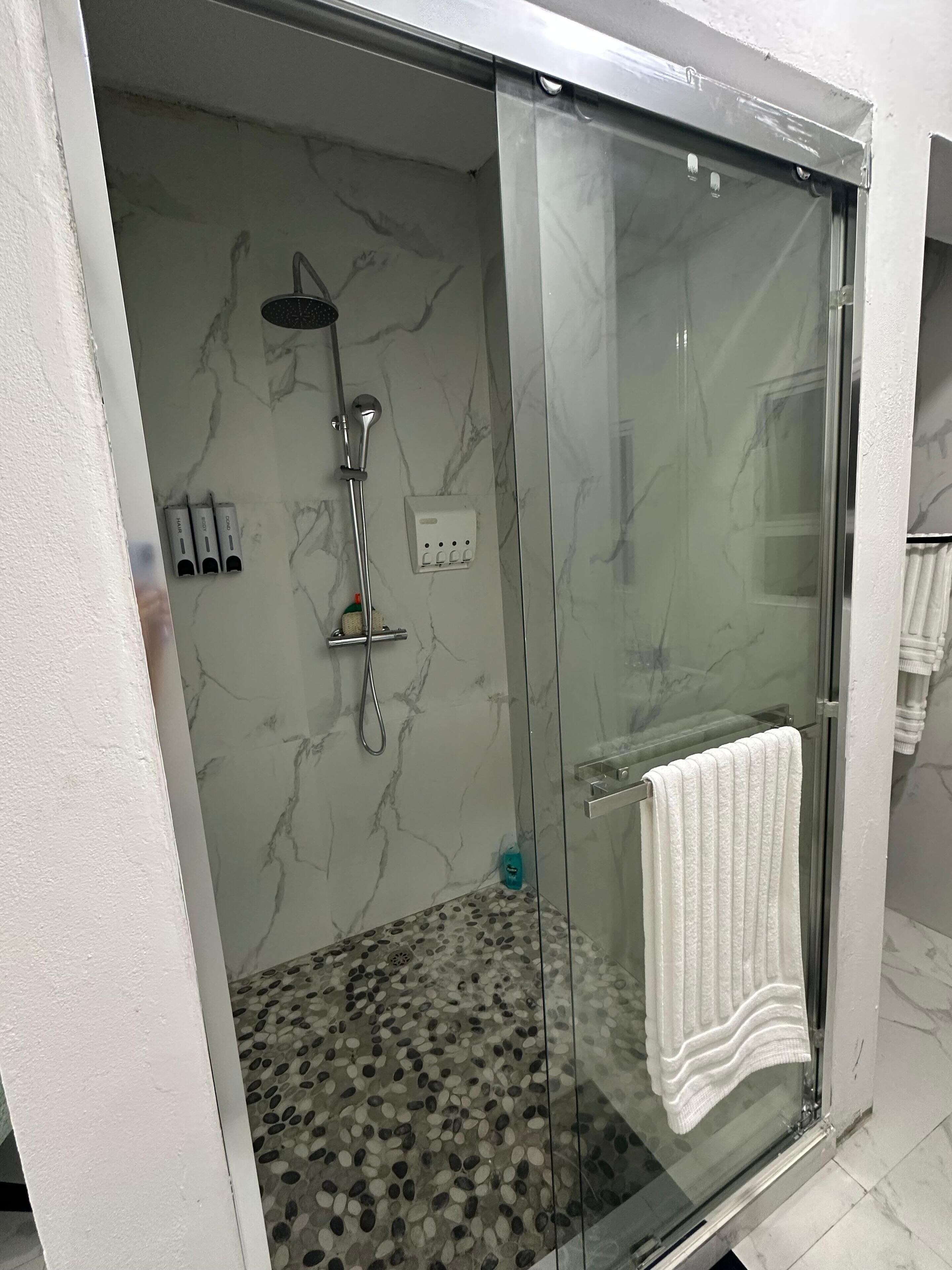 Área de chuveiro moderna com divisória de vidro, chuveiro manual e painel de controle na parede.