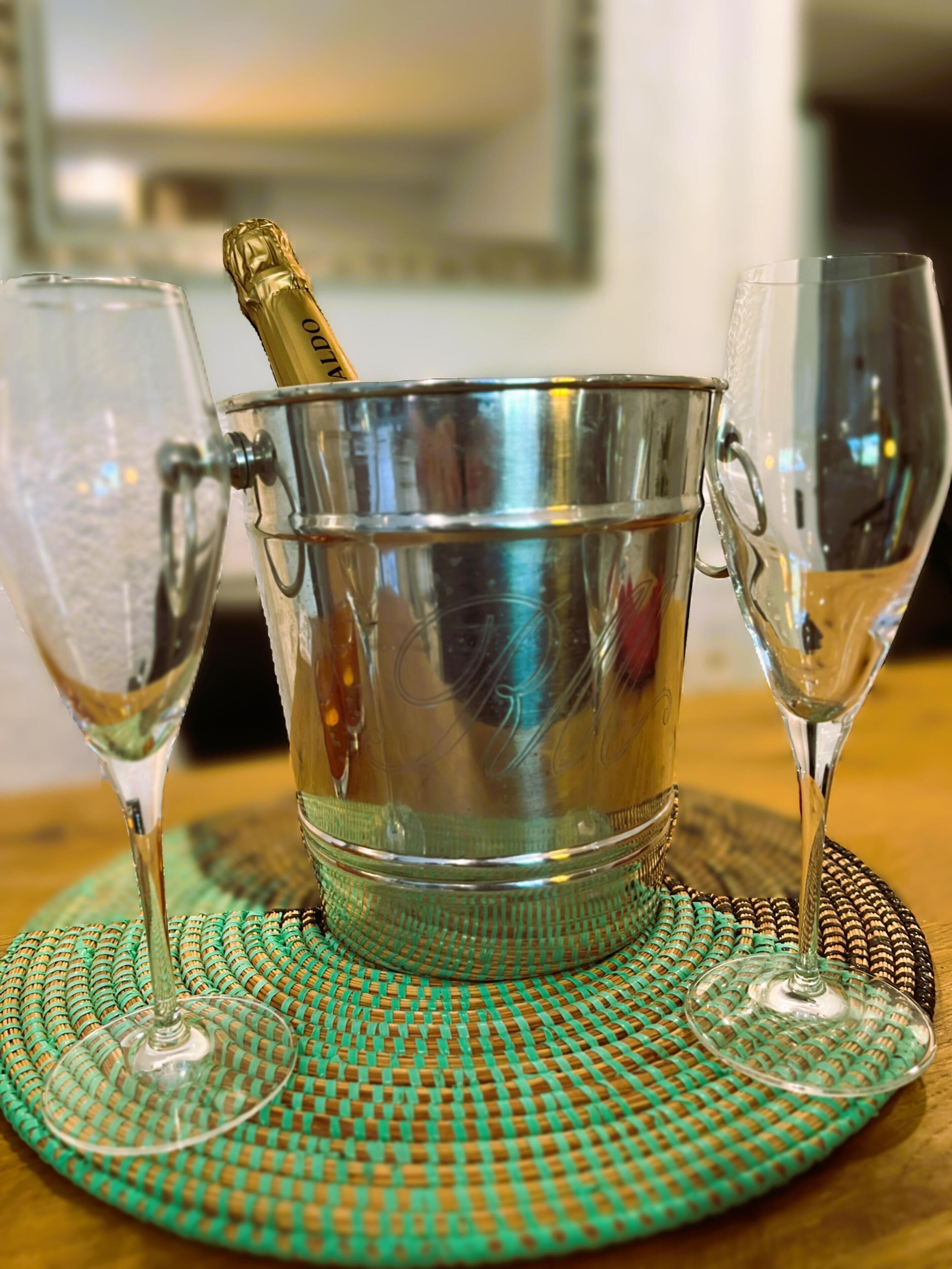 A metallic bucket with a bottle of champagne, two empty glasses, and a woven placemat on a table.