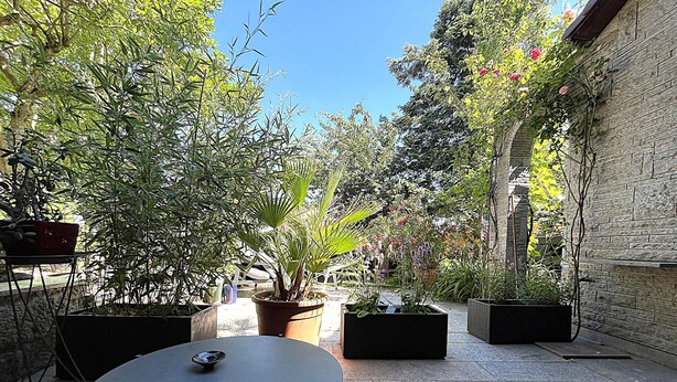 Eine Terrasse mit rundem Tisch und Stühlen, umgeben von Topfpflanzen und einer Backsteinmauer.