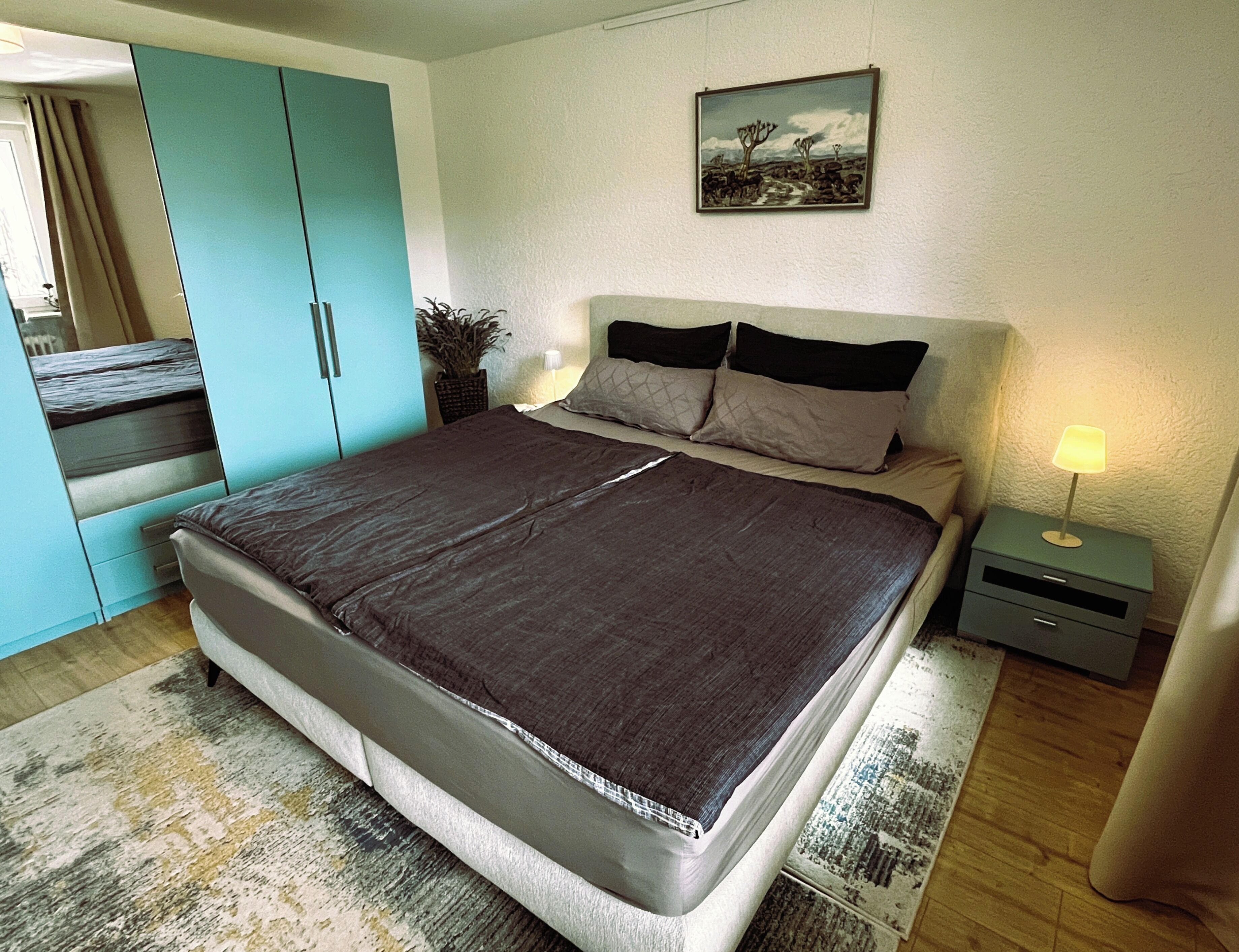 A bedroom with a bed, a nightstand, a lamp, a painting on the wall, and a wardrobe.