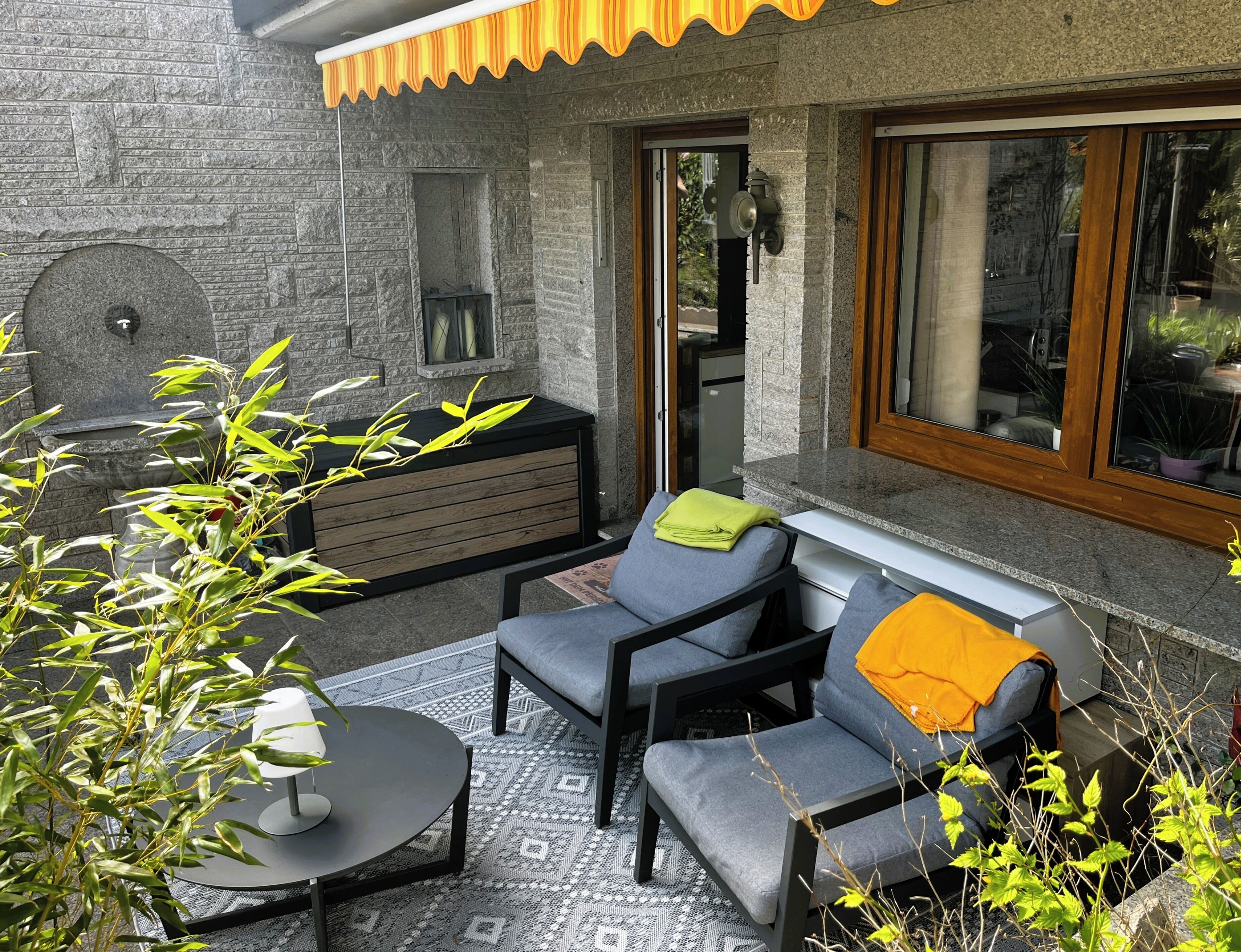 A patio area with a stone wall, a built-in bench, a grey sofa, a small round table, and a window with a view of the interior.