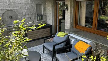 A patio area with a stone wall, a built-in bench, a grey sofa, a small round table, and a window with a view of the interior.