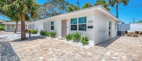 A single-story house with a white exterior, numbered 1137, featuring a front porch with a table and chairs, surrounded by a paved driveway and small shrubs.