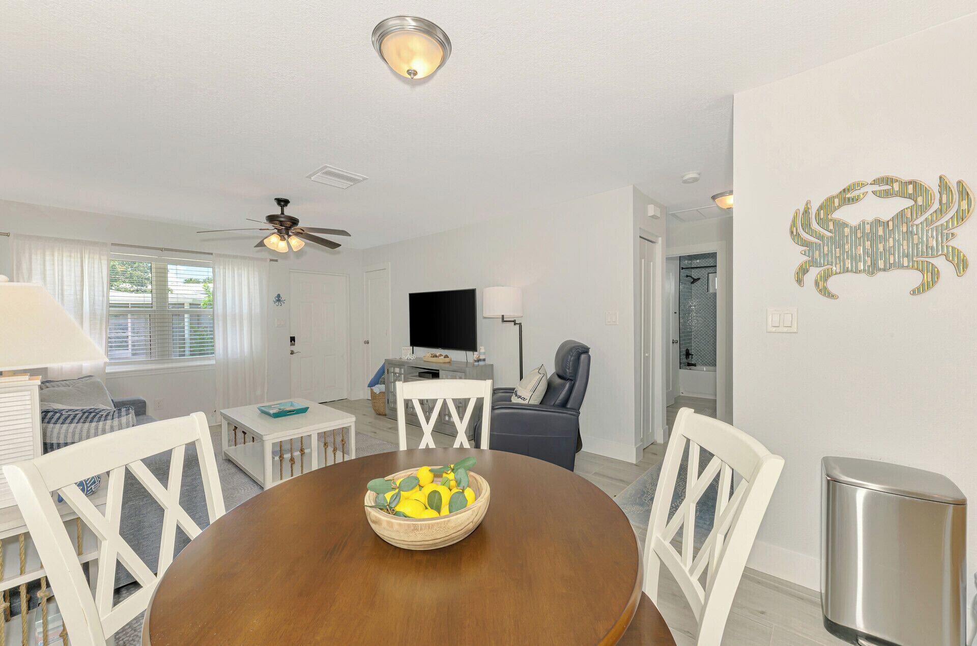 A dining area with a wooden table, white chairs, and a bowl of fruit. There is a ceiling fan, a television, and a bathroom visible in the background.
