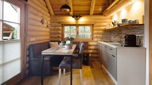 A log cabin interior with a dining table, bench seating, a kitchen area with a sink and stove, and a window with a view of greenery. - Chalet by the Lake – Vogtland Chalet: Cozy Wooden Cabin for Couples (Bad Brambach)