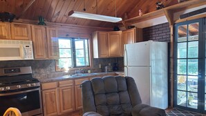 A kitchen with wooden cabinets, a microwave, stove, and refrigerator. There is a chair and a rug near the door.